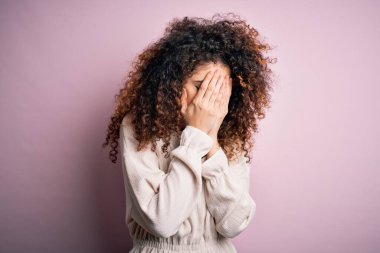 Kıvırcık saçlı ve piercingli güzel bir kadın pembe arka planda sıradan bir süveter giyiyor. Ağlarken suratını ellerle kapatıyor. Depresyon kavramı.