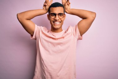 Yakışıklı Afro-Amerikalı adam pembe arka planda günlük tişört ve gözlük takıyor. Komik ve çılgın pozlar veriyor. Parmakları kafasında tavşan kulağı gibi, neşeli gülümsüyor.