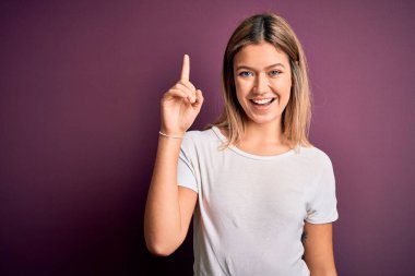 Genç, güzel, sarışın bir kadın mor izole edilmiş arka plan üzerine gündelik beyaz tişört giyip başarılı bir fikri işaret ediyor. Heyecanlı ve mutlu. Bir numara..