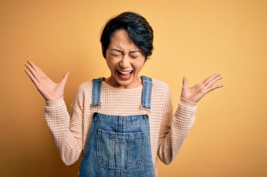 İzole edilmiş sarı arka plan üzerine sıradan kot pantolon giyen genç, güzel Asyalı kız başarı için deli ve çılgınlığını kollarını kaldırarak ve gözlerini kapatarak heyecanla bağırarak kutluyor. Kazanan konsept