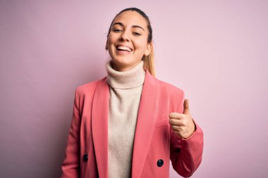 Mavi gözlü, zarif pembe ceketli, güzel sarışın iş kadını mutlu baş parmaklarıyla el hareketi yapıyor. Kameraya bakıp başarıyı gösteren ifade onaylanıyor.