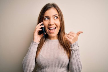 Akıllı telefondan konuşan genç bir kadın izole bir arka planda işaret ediyor ve gülümseyen bir yüzle baş parmağıyla gösteriyor.