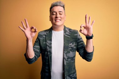 Sarı izole edilmiş arka planda duran iş ceketi giyen genç, yakışıklı, modern bir adam rahatla ve parmaklarıyla meditasyon hareketi yaparken gözleri kapalı gülümse. Yoga konsepti.