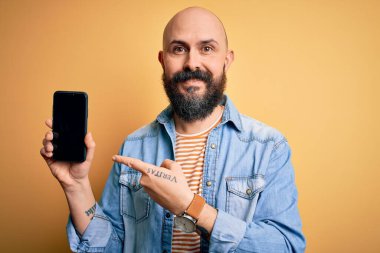 Sakallı yakışıklı kel adam. Akıllı telefon tutuyor. Sarı arka planda ekranı gösteriyor.