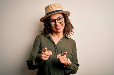 Gözlüklü ve şapkalı orta yaşlı esmer kadın izole edilmiş beyaz arka planda durmuş mutlu ve komik yüzlü parmaklarını kameraya doğrultuyor. İyi enerji ve titreşimler.