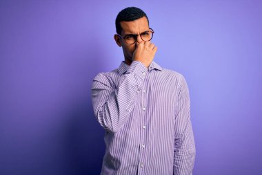 Çizgili tişört ve gözlük takmış yakışıklı Afro-Amerikan bir adam mor arka planda kötü ve iğrenç bir koku alıyor, nefesini burnunda tutuyor. Kötü kokuyor.