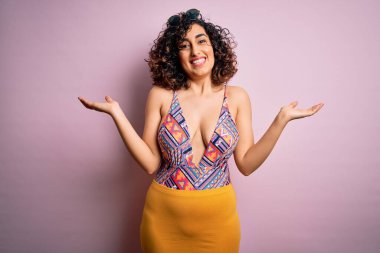 Genç, güzel bir Arap kadın, mayo ve pembe arka planda güneş gözlüğü takarak tatilde. Gülümsüyor, avuçlarını açıyor, sunuyor, reklam yapıyor, karşılaştırıyor ve dengede duruyor.