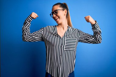 Mavi gözlü, çizgili gömlekli ve mavi arka planda gözlüklü güzel sarışın kadın gururla gülümseyen kol kaslarını gösteriyor. Fitness konsepti.