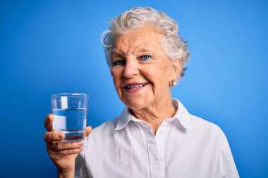 Kıdemli güzel kadın mavi arka planda duran bir bardak su içiyor. Mutlu bir yüzü var ve gülümsüyor. Kendine güvenen bir gülümsemeyle dişlerini gösteriyor.