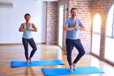 Orta yaşlı güzel sporcu çift mutlu gülümsüyor. Yüzünde gülümsemeyle ayakta dikilip, spor salonunda ağaç pozu vererek yoga yapıyor.