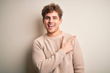 Sarışın, kıvırcık saçlı, beyaz arka planda günlük süveter giyen, yüzünde mutlu ve doğal bir ifadeyle, elleri ve parmaklarıyla gülümseyen genç, yakışıklı bir adam.