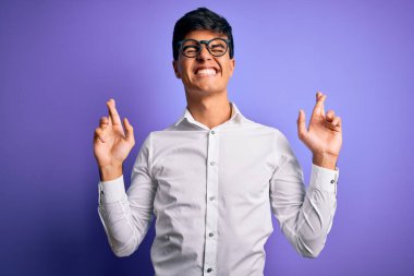 İzole edilmiş mor arka plan üzerinde gömlek ve gözlük takan genç yakışıklı iş adamı umut ve gözler kapalı gülümseyerek işaret ediyor. Şans ve batıl inanç..
