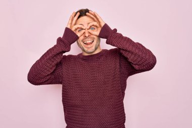 Mavi gözlü genç bir adam pembe arka planda dikilmiş normal bir süveter giyiyor. Dürbün gibi dil çıkartıyor, gözler parmaklarından bakıyor. Çılgın ifade.