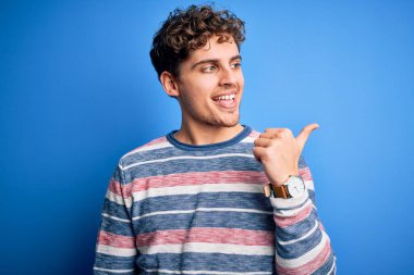 Sarışın, kıvırcık saçlı, mavi arka planda çizgili süveter giyen mutlu yüzüyle gülümseyen ve baş parmağıyla yüzünü gösteren genç bir adam..