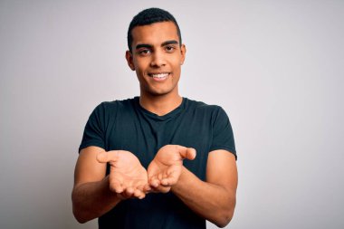 Beyaz arka planda dikilen, elleri avuçlarında gülümseyen, el hareketi yapan, sıradan tişörtler giyen genç, Afro-Amerikan bir adam. Tutun ve koruyun