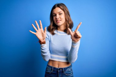 Mavi izole edilmiş arka plan üzerinde duran sıradan bir süveter giyen genç ve güzel sarışın kız yedinci parmaklarıyla işaret ederken kendinden emin ve mutlu gülümsüyor..