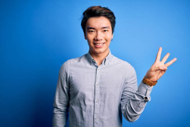 Genç, yakışıklı, sıradan bir gömlek giyen, mavi arka planda duran, üç numaralı parmaklarını gösterip gülümseyen, kendinden emin ve mutlu bir Çinli..