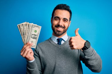 Sakallı genç yakışıklı bir adam mavi arka planda bir deste para tutuyor. Gülümsüyor, gülümsüyor. İşaret veriyor.