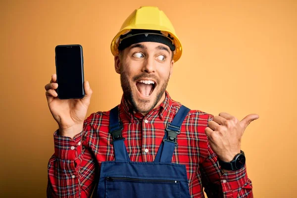 年轻的建筑工人戴着安全帽 在黄色的背景下显示智能手机屏幕 用大拇指指向侧面 笑容满面 — 图库照片