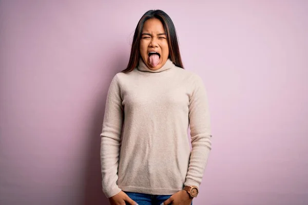 年轻美丽的亚洲女孩穿着宽松的高领毛衣 披着孤立的粉色背景 带着滑稽的表情伸出舌头 情感概念 — 图库照片