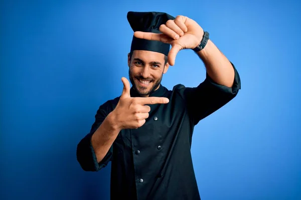 年轻英俊的厨师 留着胡子 身穿炊具制服 头戴蓝色背景的帽子 笑容满面 手指手画脚 创意和摄影概念 — 图库照片