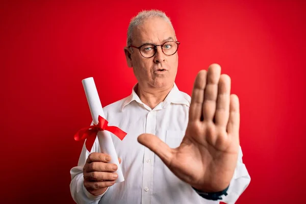 Middle age hoary student man wearing glasses holding university graduated degree diploma with open hand doing stop sign with serious and confident expression, defense gesture