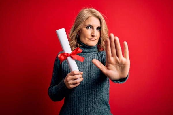 Middle age beautiful blonde student woman holding university graduate diploma with open hand doing stop sign with serious and confident expression, defense gesture