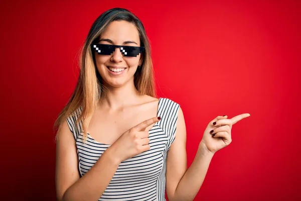 Young beautiful brunette woman wearing funny thug life sunglasses over red background smiling and looking at the camera pointing with two hands and fingers to the side.