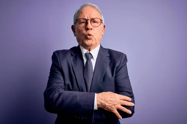 Grey haired senior business man wearing glasses and elegant suit and tie over purple background shaking and freezing for winter cold with sad and shock expression on face