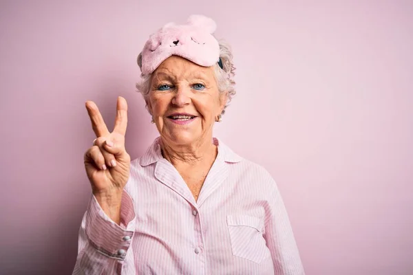 年长而漂亮的女人 戴着睡眠面具 披着睡衣 披着孤立的粉色背景 带着第二指尖 满面笑容 自信而快乐 — 图库照片