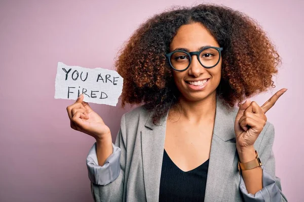 Young african american woman with afro hair holding paper with you are fired message very happy pointing with hand and finger to the side