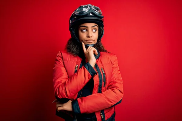 アフリカ系アメリカ人の若いオートバイ愛好家の少女は 赤い背景の上に元ヘルメットと眼鏡を着用し あごの質問 集中的な表現について考えて手をつないでいる 思慮深い顔で微笑んだ 疑いの概念 — ストック写真