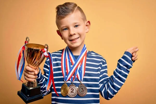 Young little caucasian kid wearing winner medals and victory award trophy over yellow background very happy pointing with hand and finger to the side