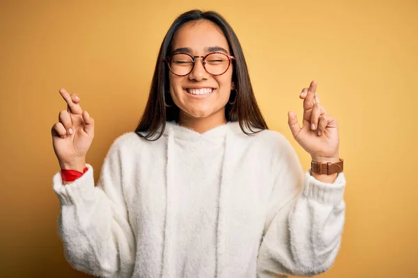 黄色の背景にカジュアルなセーターと眼鏡を身に着けている若い美しいアジアの女性ジェスチャー指は希望と目を閉じて笑顔を越えました 幸運と迷信の概念 — ストック写真