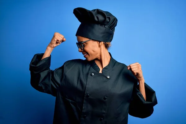 Young african american chef woman wearing cooker uniform and hat over blue background showing arms muscles smiling proud. Fitness concept.