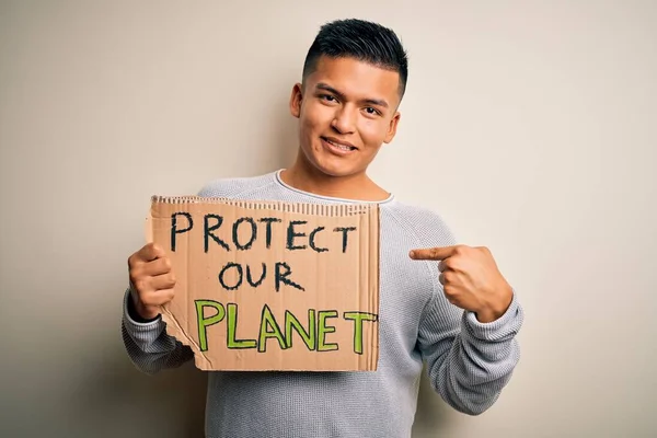 Young handsome activist latin man holding banner asking to protect our planet with surprise face pointing finger to himself