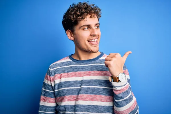 Sarışın, kıvırcık saçlı, mavi arka planda çizgili süveter giyen mutlu yüzüyle gülümseyen ve baş parmağıyla yüzünü gösteren genç bir adam..