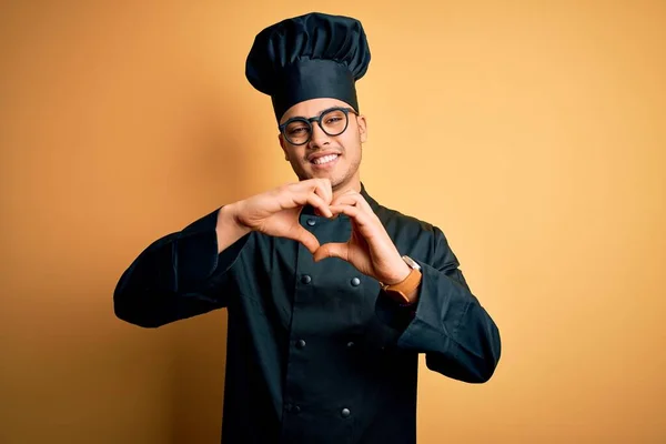 年轻的巴西厨师 身穿炊事服 头戴一顶帽子 头戴孤立的黄色背景 带着爱心微笑 手握心形符号 浪漫的概念 — 图库照片