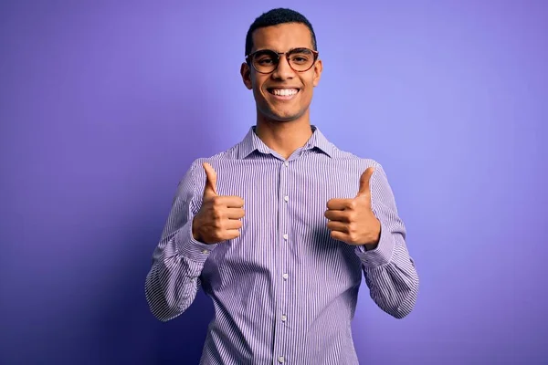 紫色の背景にストライプのシャツとメガネを身に着けているハンサムなアフリカ系アメリカ人の男性は 手で積極的なジェスチャーを行う成功サイン 親指を笑顔と幸せ 陽気な表情と勝者のジェスチャー — ストック写真