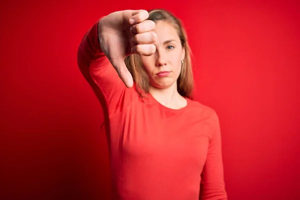 孤立した赤い背景の上に立つカジュアルなTシャツを着た若い美しいブロンドの女性は 親指を下にジェスチャーで不満と否定的な拒絶を示しています 悪い表現 — ストック写真