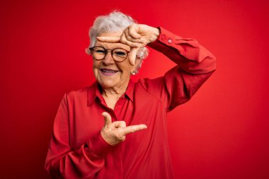 Son sınıf güzeli, gri saçlı, günlük gömlek ve gözlük takan kırmızı arka planda gülümseyen, elleri ve parmakları mutlu yüzlü bir çerçeve yapan bir kadın. Yaratıcılık ve fotoğrafçılık kavramı.