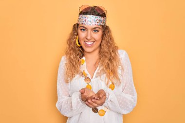 Mavi gözlü, güneş gözlüğü ve aksesuar takan, elleri avuçlarında gülümseyen ve el hareketi yapan güzel sarışın hippi kadın. Tutun ve koruyun