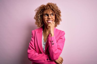 Genç Afro-Amerikalı iş kadını pembe arka planda duran gözlüklü, elleri çenesinde soru soran, düşünceli bir ifade takınıyor. Gülen ve düşünceli bir yüz. Şüphe kavramı.