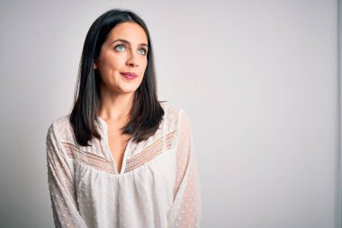 Mavi gözlü genç esmer kadın izole edilmiş beyaz arka planda günlük tişört giyiyor gülümsüyor ve başka tarafa bakıyor..