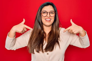 Kırmızı izole edilmiş arka planda duran gözlüklü, genç, Latin kökenli, zeki bir kadın yüzünde bir gülümsemeyle gururla ve mutlu bir şekilde kendini işaret ediyor..