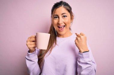 Genç, güzel esmer bir kadın izole edilmiş pembe arka planda kahve fincanı içerken gururla bağırıyor ve zaferi ve başarıyı kutluyor. Çok heyecanlı, coşkulu bir duygu.