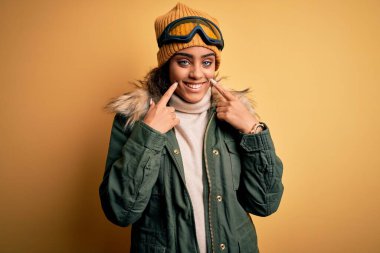 Genç Afrikalı Afro-Amerikan kayakçı kız kar sporu giyer ve kar gözlüğü takar ağzı açık gülümser, parmakları işaret eder ve neşeli bir gülümseme takınır.