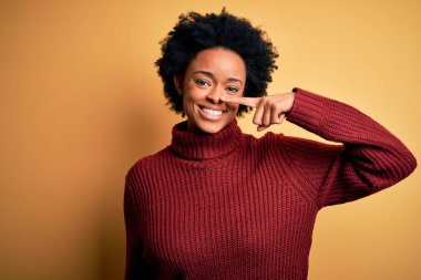 Kıvırcık saçlı, balıkçı yaka kazak giyen, el ve burnu ile işaret eden, neşeli gülümseyen güzel Afro-Amerikan kadın. Güzellik konsepti