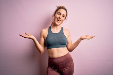 Genç ve güzel sarışın sporcu kadın spor yapıyor. Pembe arka planda spor kıyafetleri giyiyor. Gülümsüyor, yardım eli uzatıyor ve kabul ediyor..