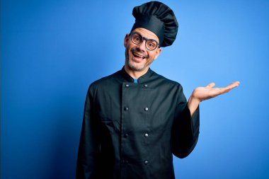 Genç, yakışıklı aşçı üniforması ve izole edilmiş mavi arka planda şapkası olan neşeli bir sunum yapan ve avuç içi kameraya bakan bir adam..